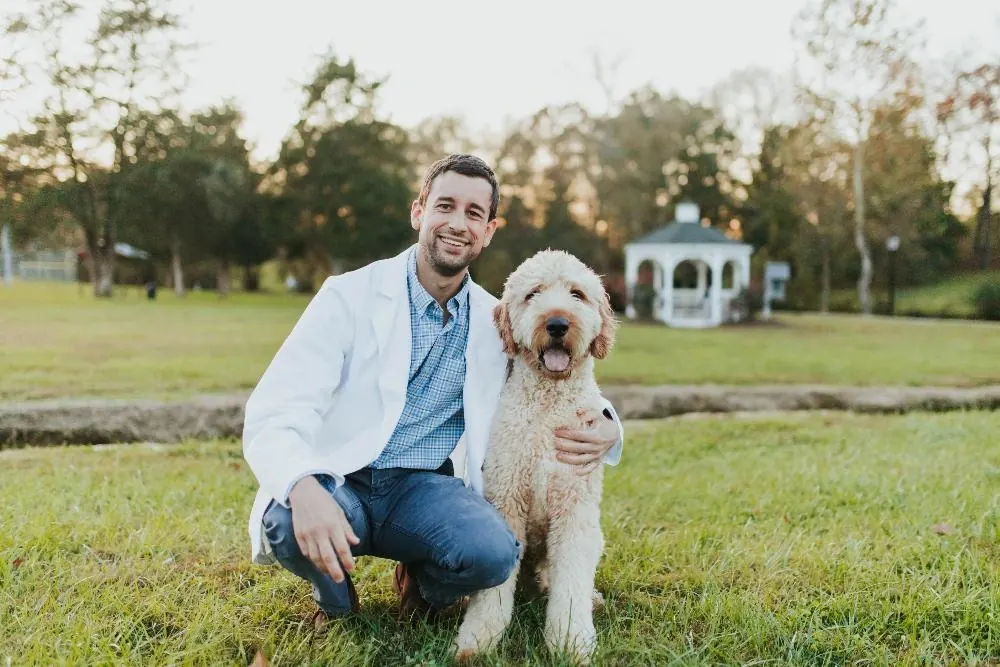 Dr. Blake Nelson kneeling on a lawn beside his goldendoodle.